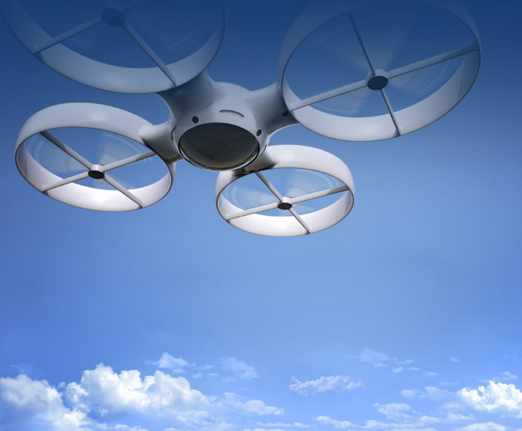 A white multi-rotor drone with ducted propellers seen from below against a bright blue sky with scattered clouds, conveying a sense of aerial surveillance and public safety technology